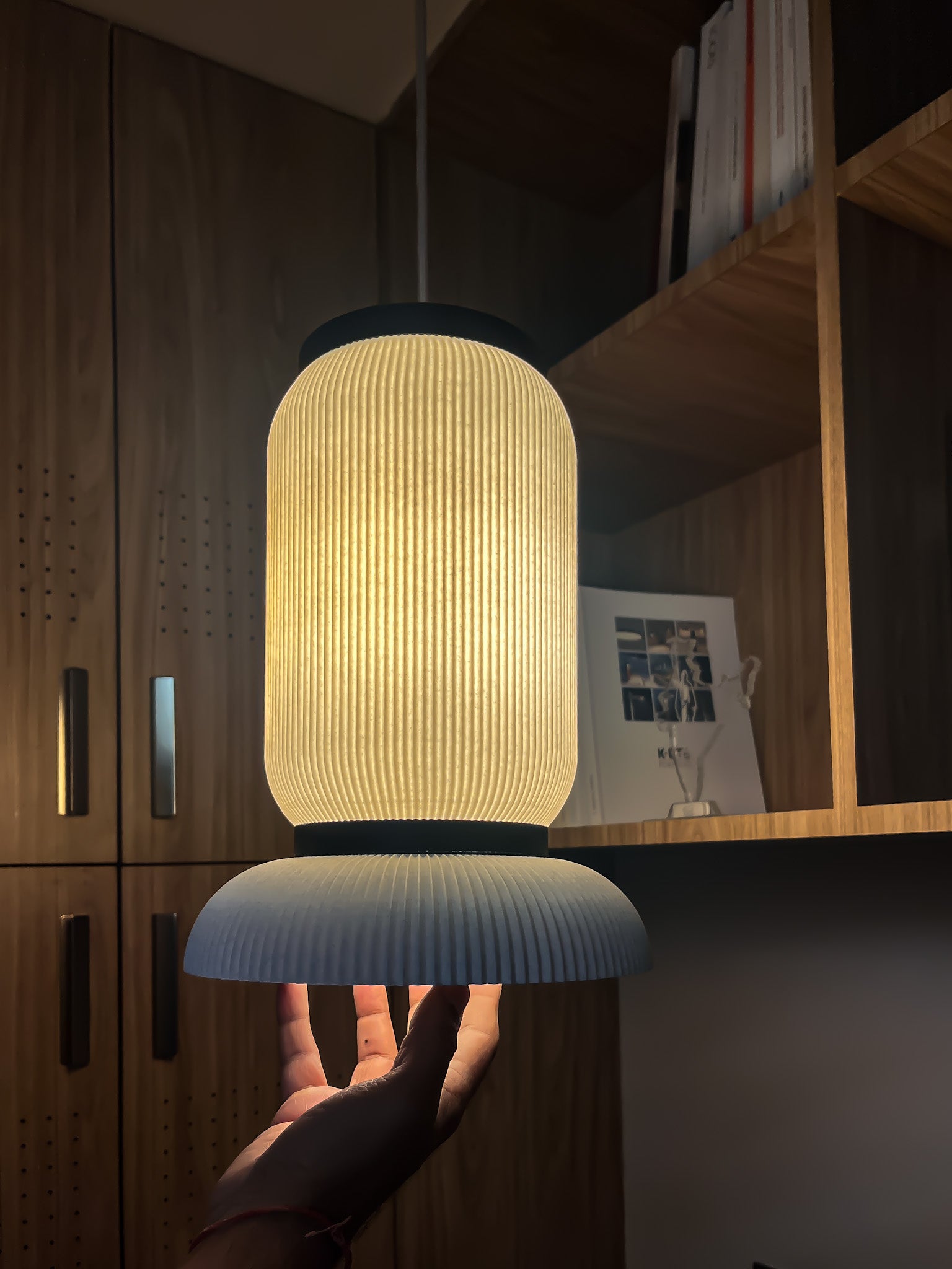 Hand holding Ganta, a textured pendant light in a room with wooden cabinets and a shelf.
